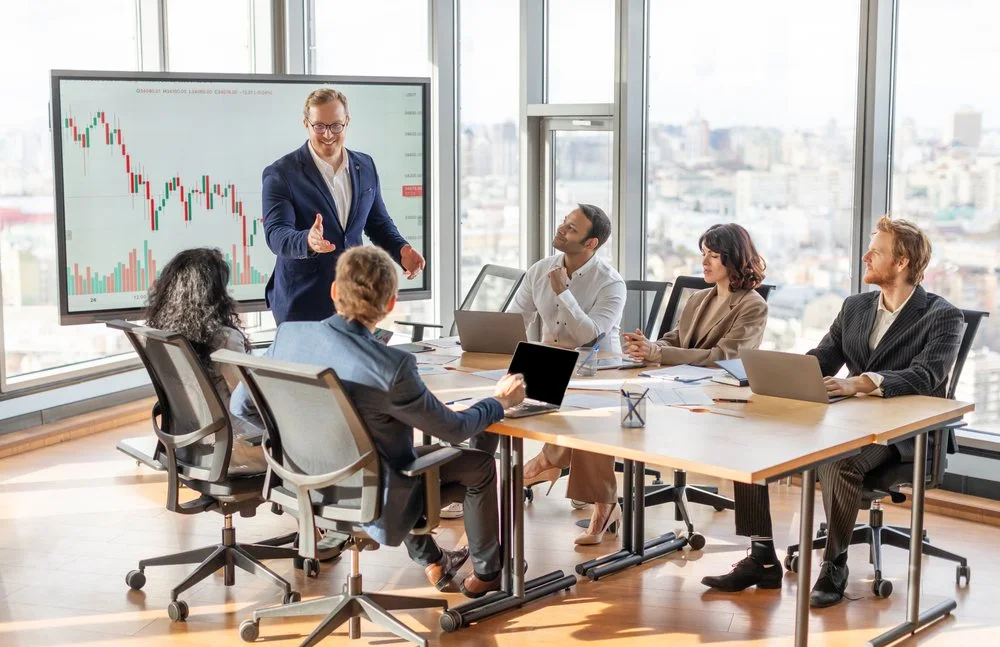 Team meeting around a conference table in front of a market chart